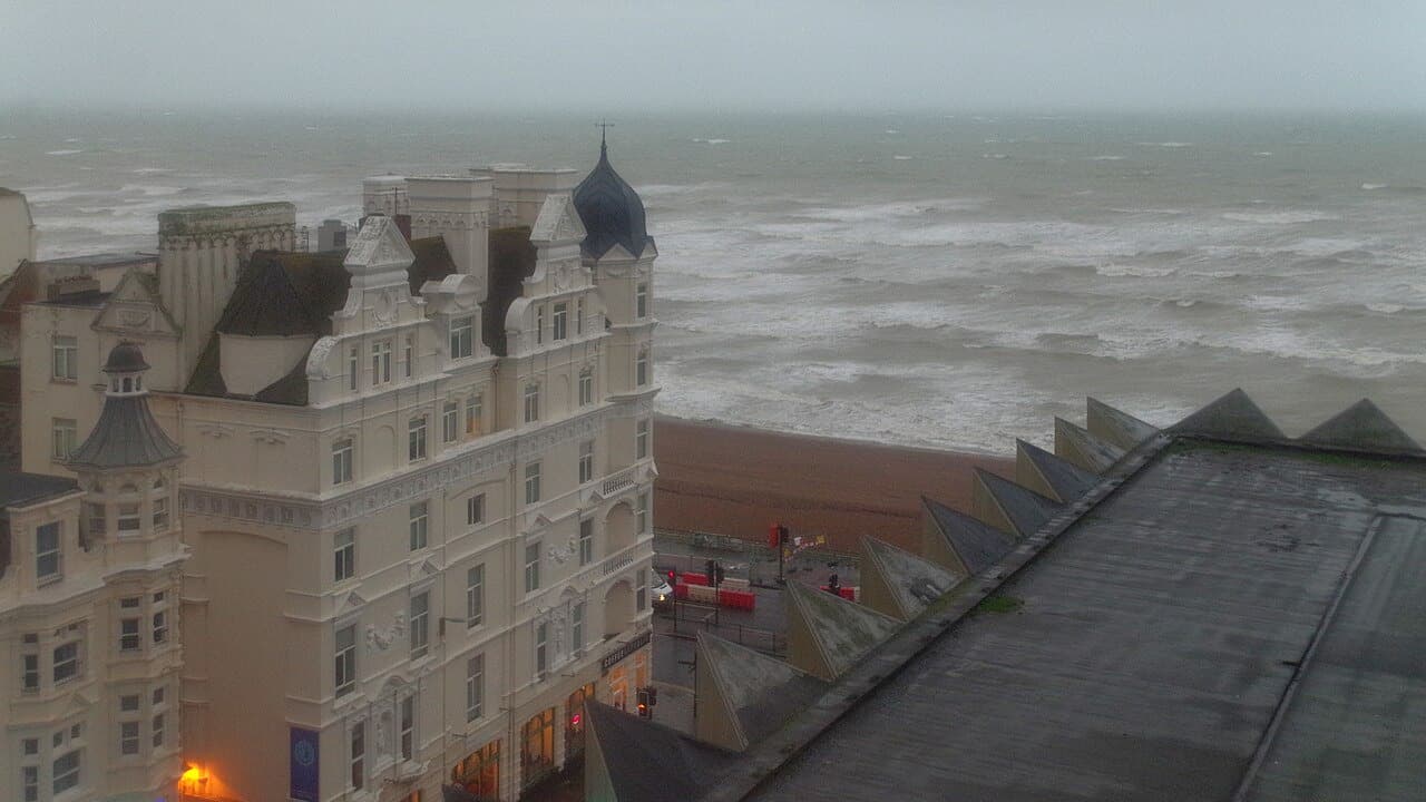 Rough grey sea and breaking waves seen from above the Brighton seafront, with a Victorian hotel on the left, on a stormy day.